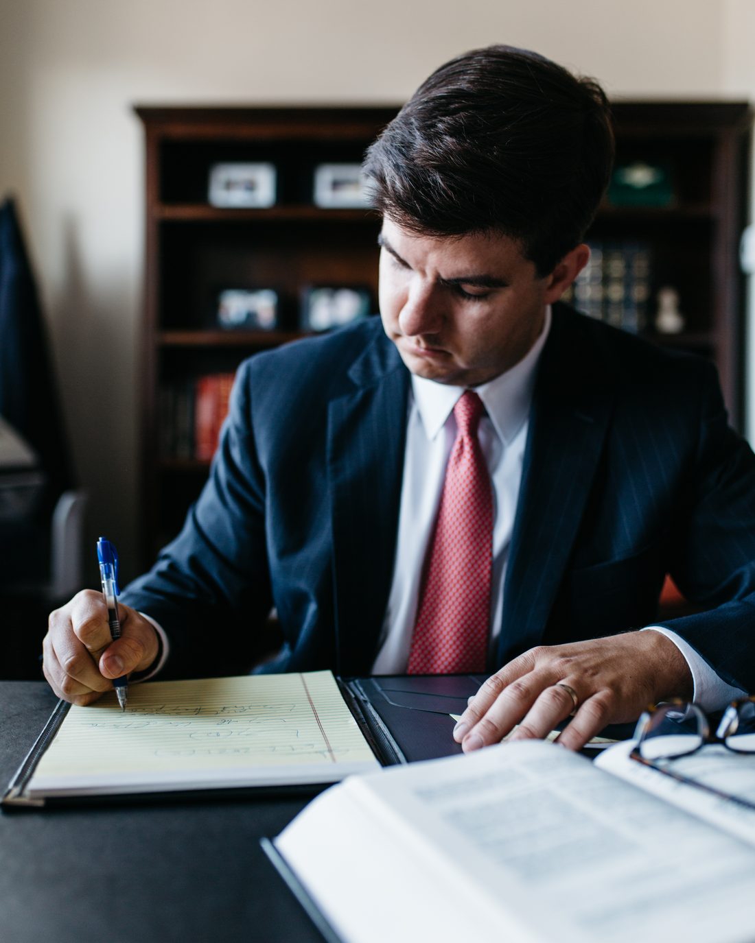 Close view of a hand writing on a yellow legal pad beside an open book and a pair of reading glasses