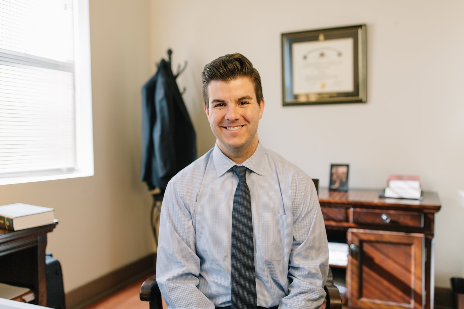 A defense lawyer smiles from a chair in a small law office, coat on the rack behind him